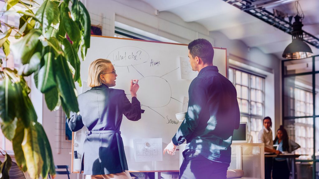 En kvinna och en man står och skriver på en vit whiteboardtavla med en grön växt i förgrunden.