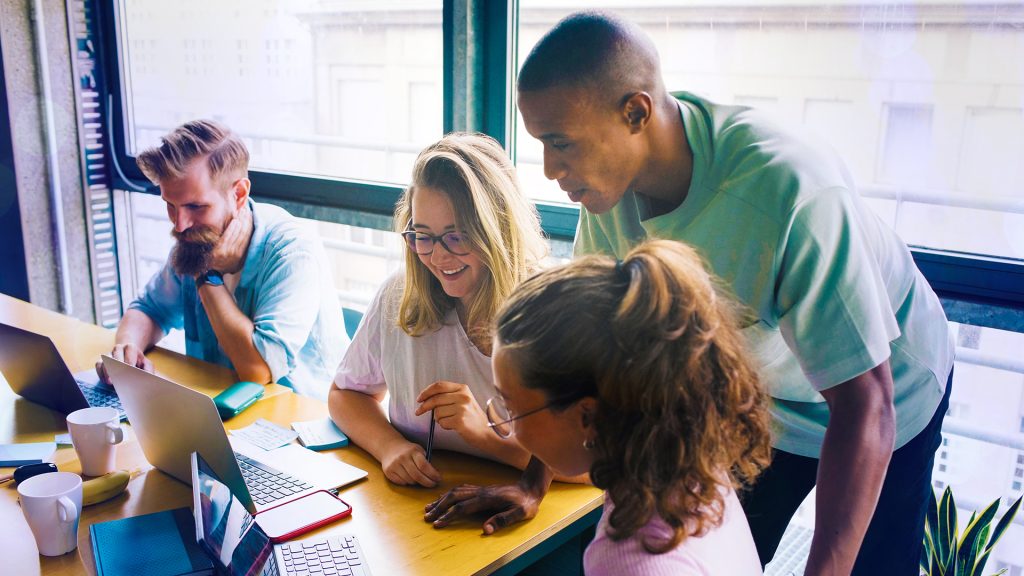Fyra personer jobbar med bärbara datorer, två är kvinnor och två är män. EN av männen står bakom en kvinna och tittar på hennes datorskärm.