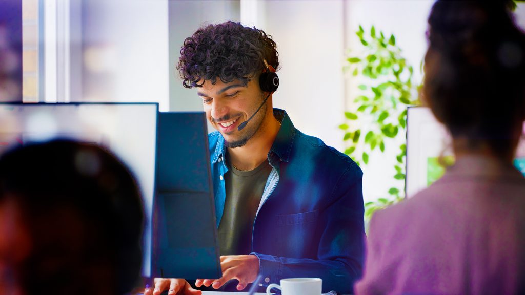 En leende man sitter med ett headset på sig och skriver på sin dator