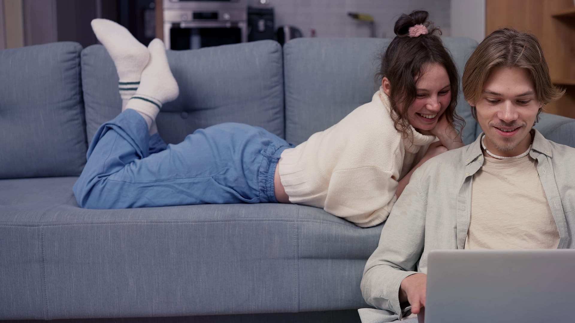 Tjej och kille i hemmiljö hon ligger på soffa och han sitter på golvet, tittar på laptopskärm och ler.