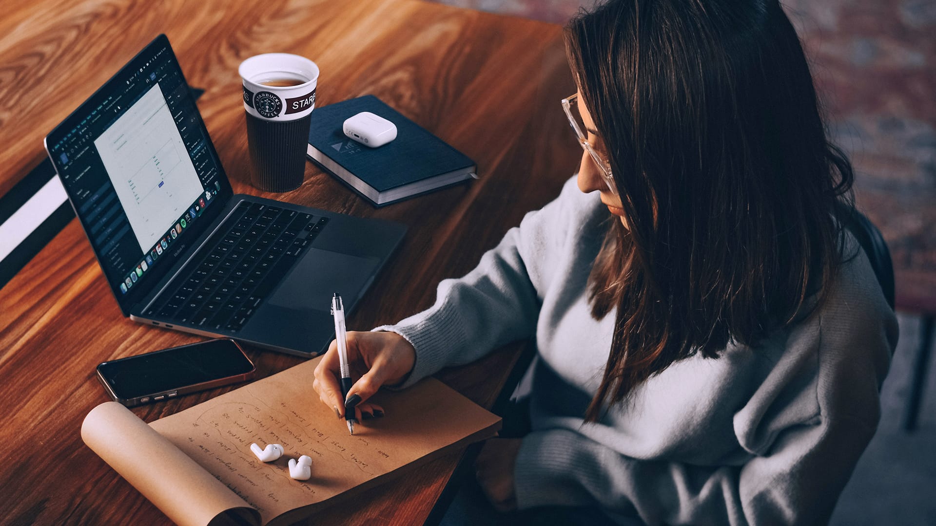 Tjej med mörkt hår fotad uppifrån, skriver på ett block framför dig med laoptop och kaffe i bakgrunden