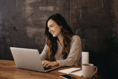 Brunhårig tjej sitter vid bord och ler med laptop framför sig.