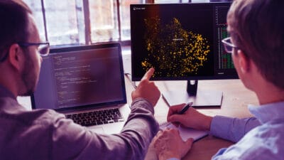Två män i kontorsmiljö fotade bakifrån som sitter med datorskärmar framför sig och diskuterar och pekar mot en av skärmarna